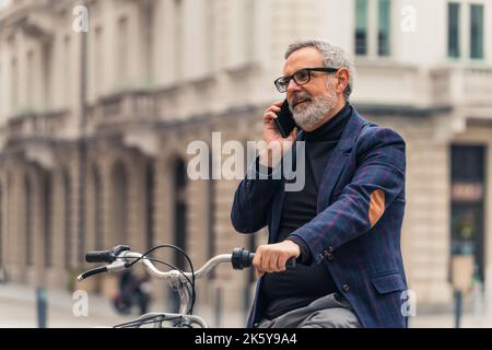 Imprenditore maschio caucasico anziano che trascorre attivamente il suo tempo libero. In bicicletta attraverso il centro della città. Uomo bearded grigio-capelli in occhiali con cornice nera, e blazer controllato parlando tramite smartphone. Foto di alta qualità Foto Stock