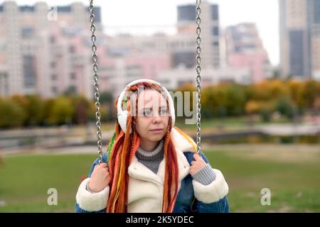 Giovane donna con lucchetti in cuffie wireless che oscillano sull'altalena, ascoltando musica nel parco cittadino. Bella femmina con capelli colorati multicolori Foto Stock