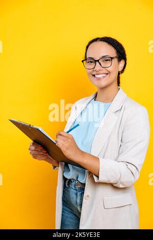 Foto verticale di giovane latino o brasiliana, in elegante abbigliamento casual, hr manager, agente immobiliare, tiene cartella con documenti, guarda fotocamera e sorriso, isolato sfondo arancione Foto Stock