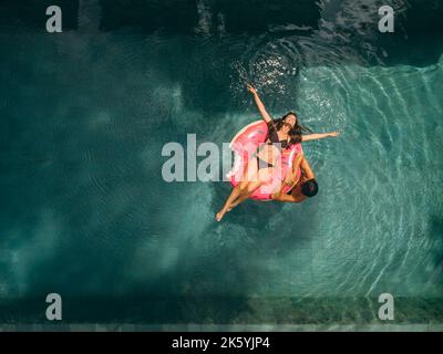 Vista aerea della coppia che si diverte nella piscina del resort. Donna sdraiata sull'anello di galleggiamento della ciambella e uomo in acqua accanto a lei. Foto Stock