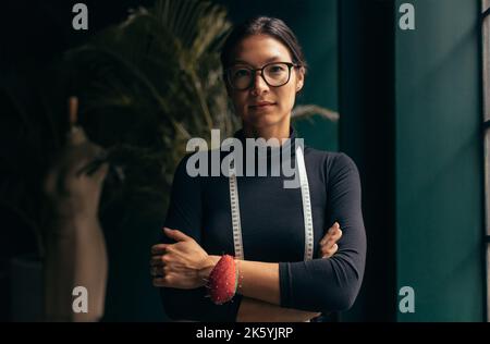 Ritratto di una stilista di moda femminile che guarda la fotocamera mentre si trova sul posto di lavoro. Donna in piedi in studio di moda con le braccia incrociate. Foto Stock