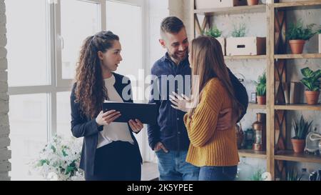 La famiglia giovane felice sta parlando con l'agente di alloggiamento all'interno del nuovo appartamento circa l'acquisto del bene immobile, hugging e gesturing emozionalmente. Concetto di casa commovente e di proprietà d'acquisto. Foto Stock