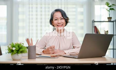 Felice e affascinante 60s donna asiatica o donna d'affari seduto alla sua scrivania, sorridendo e guardando la macchina fotografica. Foto Stock