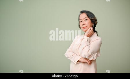 Splendida e premurosa donna asiatica di 60s anni in abiti casual, braccio attraversato, mano sul mento, guardando la macchina fotografica, in piedi isolato sul retro verde studio Foto Stock