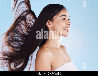 Una bella giovane donna ispanica con pelle sana e capelli lisci che soffiano nel vento sorridendo su uno sfondo blu studio. Felice misto Foto Stock