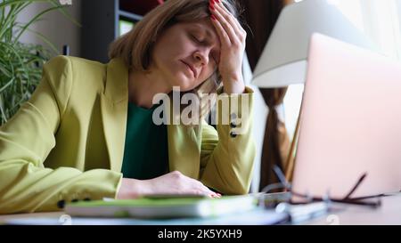 Donna sovraccarica seduta in ufficio e che tocca la testa con la mano Foto Stock