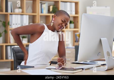 Giovane imprenditrice afroamericana che soffre di mal di schiena mentre si siede su una sedia e utilizza un computer in un ufficio al lavoro. Donna nera infelice Foto Stock