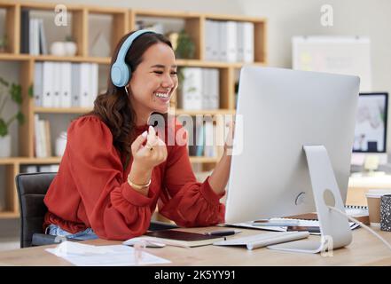 Giovane donna d'affari asiatica felice che indossa le cuffie mentre lavora su un computer in un ufficio. Una donna che ascolta solo musica, videochiama Foto Stock