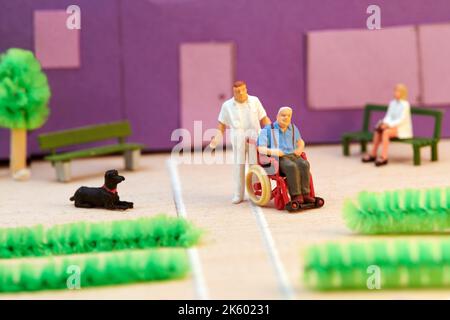 Infermiera in plastica in miniatura per aiutare il paziente anziano su sedia a rotelle di fronte all'edificio dell'ospizio Foto Stock