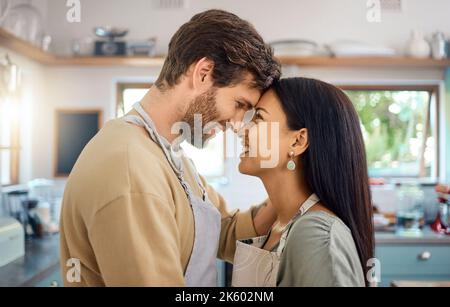 Felice coppia interrazziale toccare fronte mentre condividere romantico momento intimo a casa, indossando grembiuli e cucinare insieme. Giovane caucasico Foto Stock