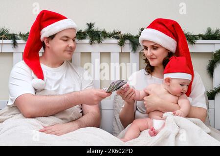 Marito dà soldi dollari a moglie con bambino sul letto decorato per natale e Capodanno, coppia di famiglia in camera da letto casa Foto Stock