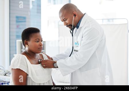 Medico maturo che usa uno stetoscopio durante una consultazione. Dottore afro-americano che ascolta il battito cardiaco dei pazienti. Donna che ha il suo petto controllato da un Foto Stock