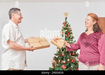 Coppia ispanica anziana che trascorre felicemente le vacanze a casa, scambiando regali di Natale vicino a un albero decorato. La gioia di celebrare insieme. Foto Stock