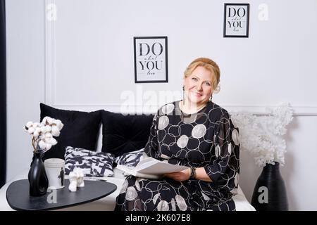 Donna anziana si siede comodamente su un divano e legge un libro Foto Stock