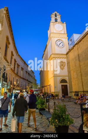 Turisti e locali passeggiando davanti alla torre dell'orologio della Cattedrale di Gallipoli su una strada acciottolata nel centro storico di Gallipoli in serata, Puglia, Italia. Foto Stock