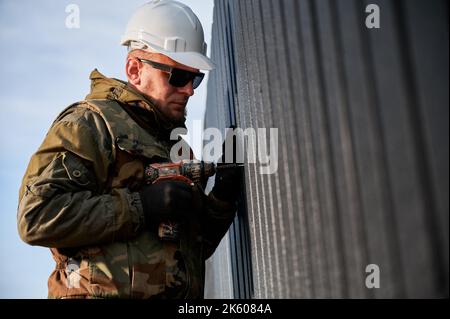 Costruttore maschio che installa nero ondulato foglio di ferro usato come facciata del cottage futuro. Uomo lavoratore edificio legno telaio casa. Carpenteria e concetto di costruzione. Foto Stock