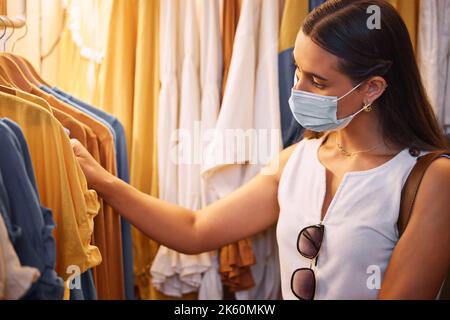 Donna che indossa una maschera durante lo shopping. Donna che sceglie un vestito in un negozio. Il virus Corona non la fermerà da una corsa allo shopping. Spendere soldi per trendy Foto Stock