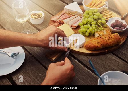 Uomo sconosciuto che prende e taglia formaggio da una varietà di spuntini su un tabellone di legno di tapas all'esterno. Pane, uva fresca e salumi preparati per il pranzo Foto Stock
