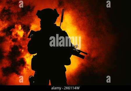 Silhouette di moderno soldato di fanteria, combattente militare d'élite in munizioni tattiche e casco, in piedi con fucile assalto in mano nel fuoco e nel fumo Foto Stock