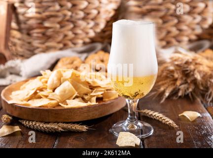 Versare la birra nel bicchiere. Spikelets di grano con una tazza di birra su sfondo di legno. Birra lager fredda. La birra artigianale forma delle onde. Freschezza e schiuma. Nelle vicinanze ci sono sottili patatine in sardo Foto Stock