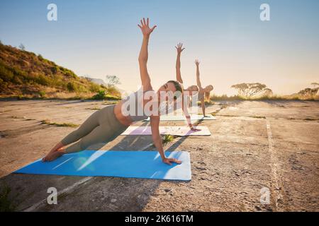 Donne di yoga a lunghezza intera che tengono la posa della tavola laterale nella pratica esterna nella natura remota. Gruppo vario di amici seri belli utilizzando tappetini e. Foto Stock