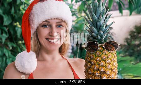 Bella bionda sorridente nel cappello di Babbo Natale guardando la macchina fotografica, tenendo l'ananas indossando occhiali da sole sullo sfondo di piante tropicali verdi. Concetto Foto Stock