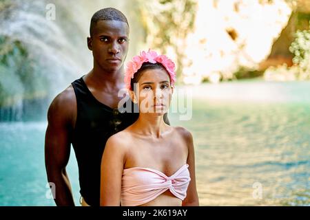 Uomo nero in abiti casual abbracciando ragazza ispanica con corona floreale su sfondo sfocato di lago pulito e scogliera soleggiato nel fine settimana Foto Stock