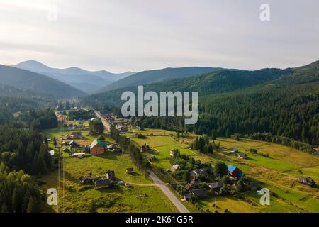 Vista aerea di mattina di nebbia luminosa sopra le piccole case rurali tra cime scure con alberi di foresta di montagna all'alba di autunno. Bellissimo scenario di selvaggio Foto Stock