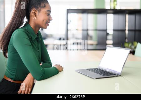 Giovane studentessa afro-americana che siede alla scrivania in un moderno spazio di coworking di fronte al computer portatile, guardando lo schermo bianco del computer. Vista posteriore a spalla Foto Stock