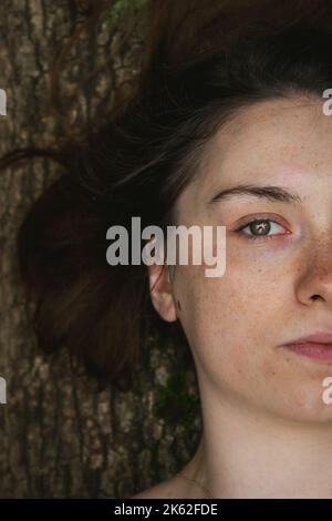 Metà del volto di una bella giovane donna con gli occhi di nocciolo, sdraiata su un tronco di albero in una foresta Foto Stock