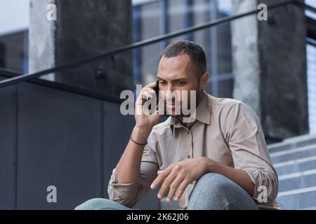 Uomo depresso seduto sulle scale dell'edificio dell'ufficio, frustrato parlando al telefono, rovesciato e licenziato primo piano di lavoro in camicia casual. Foto Stock