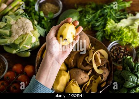 Una donna tiene in mano una patata sbucciata cruda. Sul tavolo si trovano varie verdure fresche. Cucina vegetariana cibo. Foto Stock