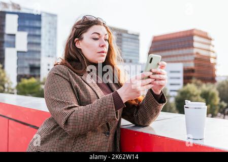 Donna in outerwear con capelli lunghi appoggiati al bordo e fotografare una tazza di caffè da asporto senza sprechi di giorno sulla strada cittadina Foto Stock