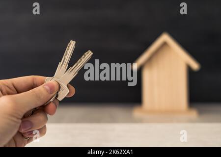 Una mano della persona che tiene le chiavi alla loro nuova proprietà domestica con la piccola casa di legno sullo sfondo. Foto Stock