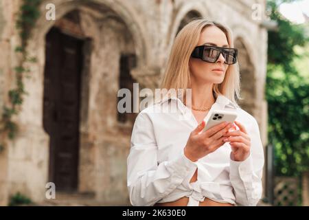 Donna fiduciosa che texting su smartphone mentre in piedi per strada, elegante Businesswoman indossare occhiali da sole utilizzando il telefono cellulare e guardando lontano. Foto Stock