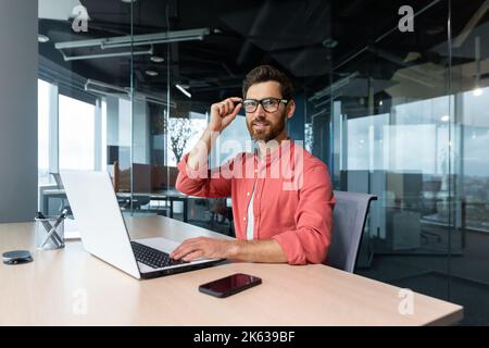 Ritratto di un uomo d'affari maturo freelancer startup, uomo bearded sorridendo e guardando la macchina fotografica, il proprietario di affari che lavora all'interno di un moderno edificio di uffici indossando camicia rossa e occhiali Foto Stock