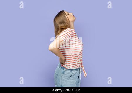 Vista laterale di una donna bionda che indossa una T-shirt a strisce che tocca la schiena e grida di dolore, che soffre di mal di schiena, che ha fastidio muscolare lombare inferiore. Studio al coperto isolato su sfondo viola. Foto Stock