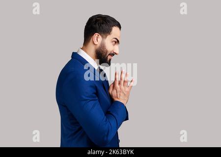 Vista laterale dell'uomo bearded astuto che stringe le mani e che progetta il pungolo ingannevole o scheming malvagio, barare con il sorriso silly, portando il vestito ufficiale di stile. Studio in interni isolato su sfondo grigio. Foto Stock