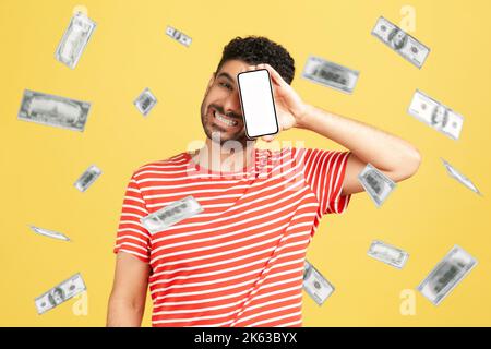 Investimento in linea, vincitore overjoyed uomo ricco e guadagnare soldi. Un ragazzo soddisfatto che copriva l'occhio con un display mobile vuoto e gioisce della sua ricchezza. Studio al coperto isolato su sfondo giallo. Foto Stock