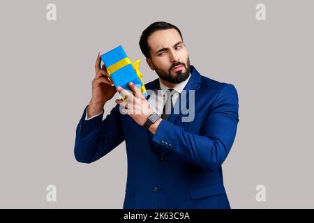 Ritratto di curioso uomo d'affari bearded tenendo e scuotendo confezionato presente scatola, essendo interessato che cosa all'interno, indossando tuta ufficiale di stile. Studio in interni isolato su sfondo grigio. Foto Stock