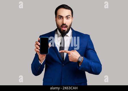 Uomo bearded che punta il dito contro lo smartphone con schermo vuoto, guardando la fotocamera con shock, spazio libero per la pubblicità, indossando tuta di stile ufficiale. Studio in interni isolato su sfondo grigio. Foto Stock