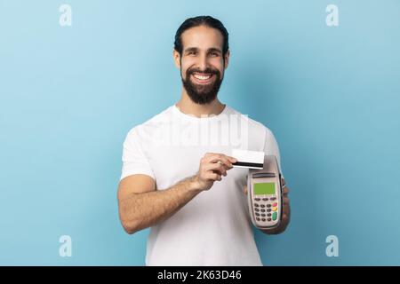 Ritratto di uomo sorridente con barba che indossa una T-shirt bianca che tiene in mano il terminale di pagamento POS, andando a effettuare il pagamento con carta, acquisti senza contanti. Studio in interni isolato su sfondo blu. Foto Stock