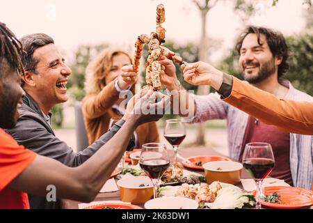 Foto all'aperto di amici multietnici che si godono una festa barbecue in giardino, seduti al tavolo e allietando con spiedini di carne. - amicizia, cibo Foto Stock