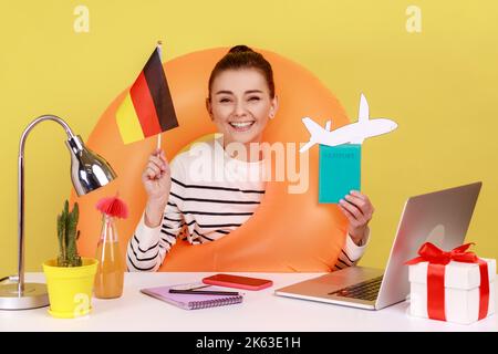 Ritratto di una donna molto contenta, responsabile ufficio con anello di gomma in possesso di passaporto e bandiera tedesca, felice di viaggiare in Germania. Studio interno girato isolato su sfondo giallo. Foto Stock