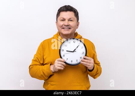 Uomo positivo che indossa una camicia blu casual che mostra tenendo grande orologio da parete, guardando la macchina fotografica con sorriso toothy, gestione del tempo, indossando felpa con cappuccio stile urbano. Studio in interni isolato su sfondo bianco Foto Stock