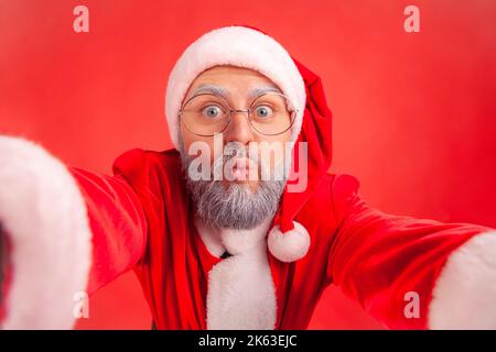 Felice uomo anziano positivo con barba grigia indossando il costume di babbo natale facendo selfie POV, guardando la macchina fotografica con gesto di bacio, espressione divertente. Studio in interni isolato su sfondo rosso. Foto Stock