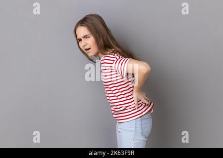 Vista laterale ritratto della bambina che indossa una T-shirt a righe urlando in dolore acuto e tenendo mal di schiena, rischio di malattie da calcoli renali, nervo pizzicato. Studio in interni isolato su sfondo grigio. Foto Stock