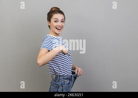 Attraente donna che indossa T-shirt a righe hanno willpower a perdere peso, puntando a vita sottile e puntando il dito su pantaloni grandi, la concezione della dieta. Studio in interni isolato su sfondo grigio. Foto Stock