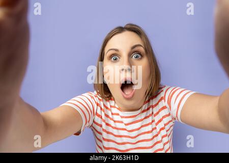 Ritratto di donna scioccata in T-shirt a righe scattando foto selfie, punto di vista, guardando la fotocamera con bocca aperta, facendo autoritratto anteriore. Studio al coperto isolato su sfondo viola. Foto Stock