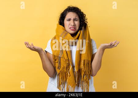 Ritratto di donna malata malsana con capelli ondulati scuri avvolti in sciarpa calda, essendo impotente, piangendo che soffre di sintomi influenzali, si sente male. Studio al coperto isolato su sfondo giallo. Foto Stock